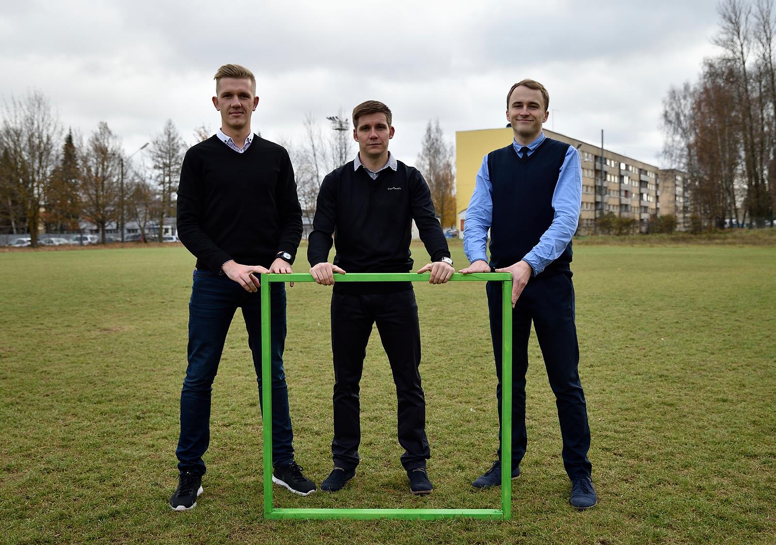 Three men standing on stadium grass while holding onto a wooden green square meter.