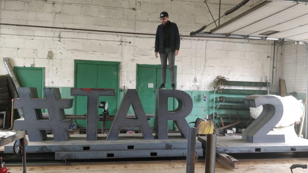 A photo of Kalle Paas standing on top of an unpainted 3D metal letter R, that is part of the #TARTU2024 landmark that's being made.