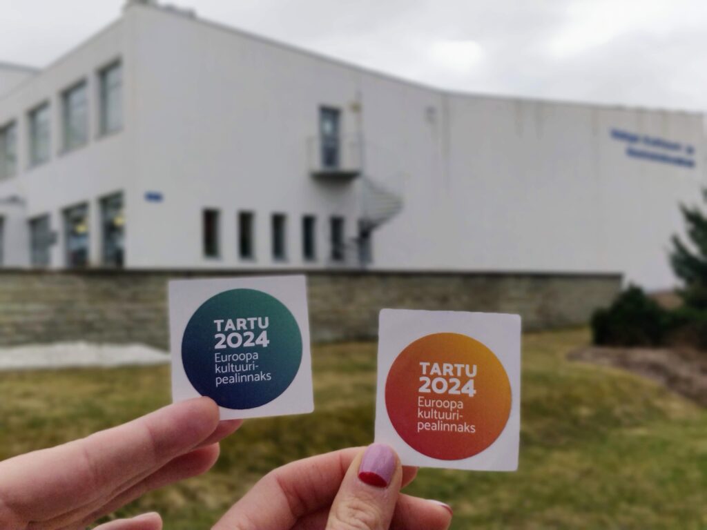 A photo of two hands holding two Tartu 2024 stickers in front of a white building